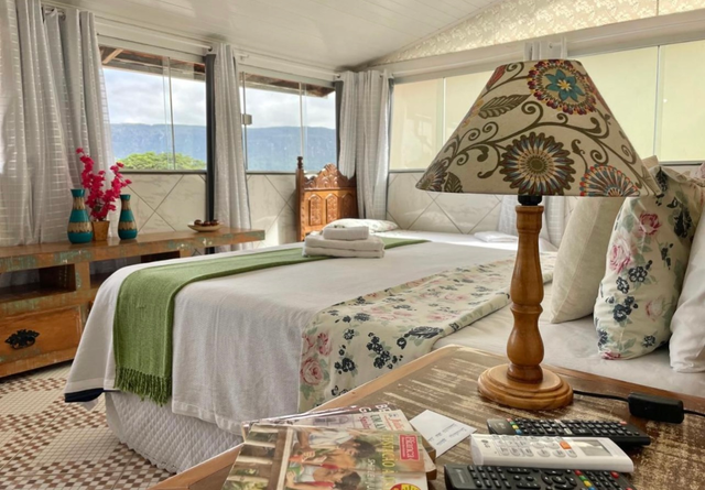 Quarto aconchegante com decoração floral, cama arrumada, abajur de madeira e amplas janelas com vista para as montanhas.