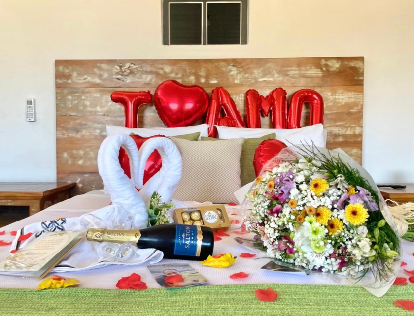 Quarto romântico decorado com balões em formato de coração, champanhe, chocolates e um buquê de flores coloridas na cama.
