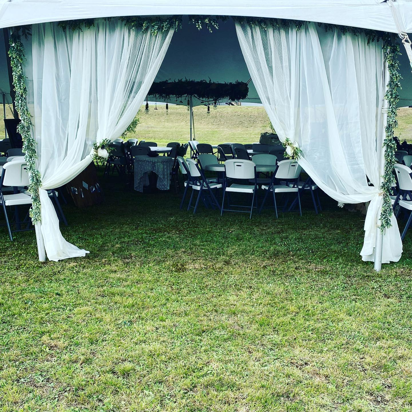 White-draped outdoor canopy with tables and chairs set on a grassy field under an overcast sky