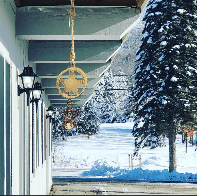 Snowy porch with hanging lanterns and a wreath-like decoration beside a white building and evergreen trees.