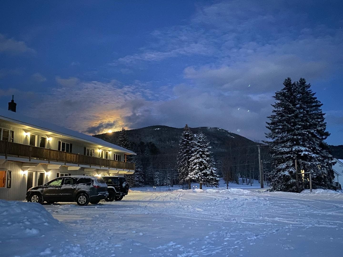 Snowy motel parking lot at dusk with lit rooms, parked SUVs, and pine trees under a cloudy blue sky
