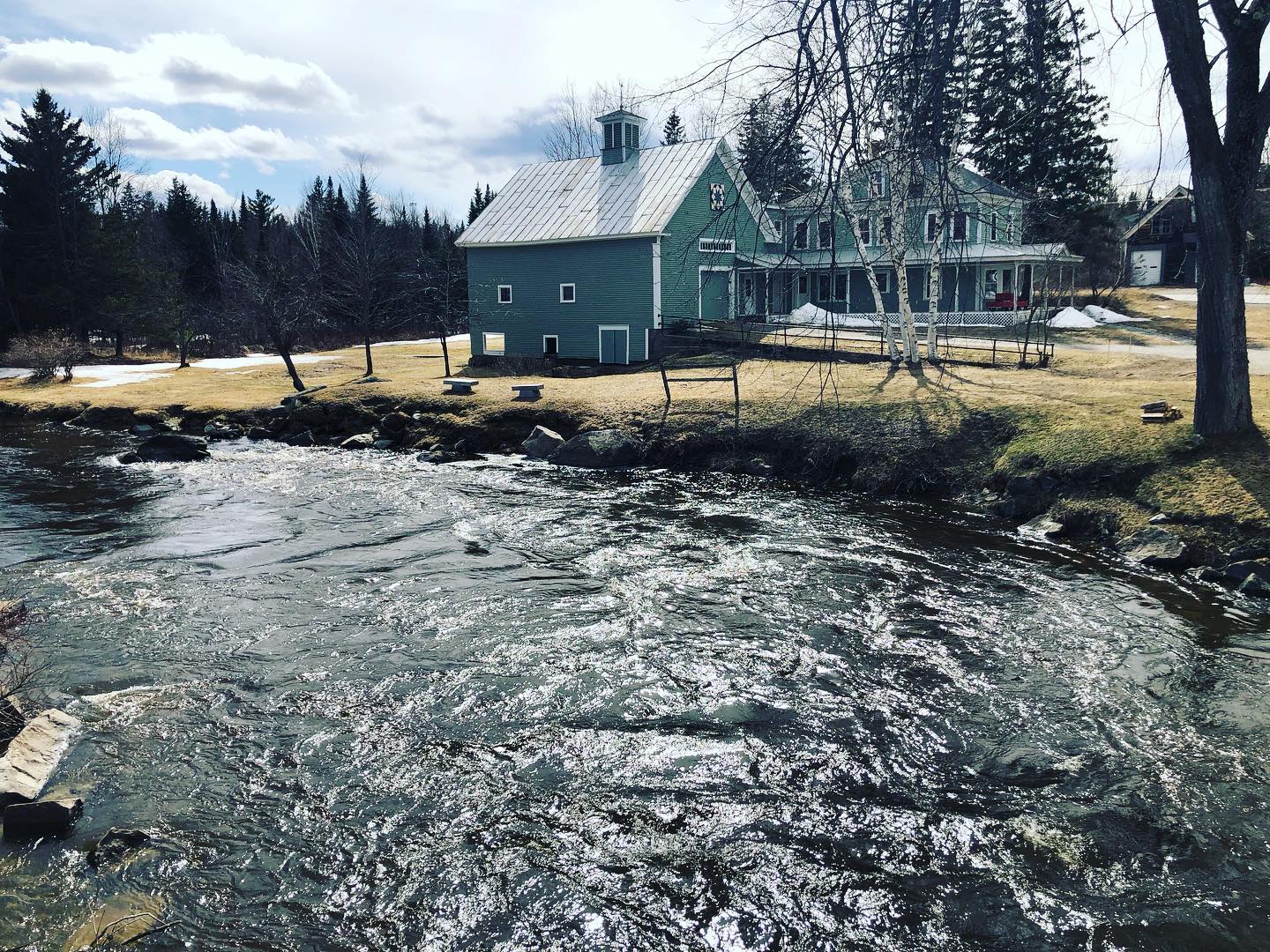 Rushing stream beside a green house and barn under leafless trees in a rural winter landscape