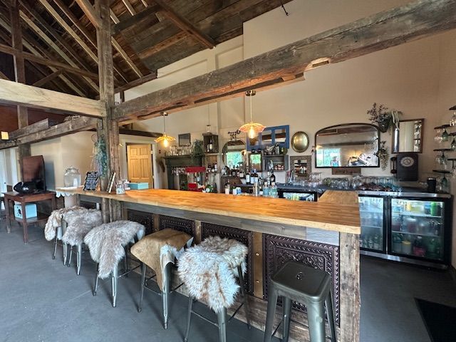 Rustic bar interior with wooden counter, furry stools, shelves of bottles, and exposed beams