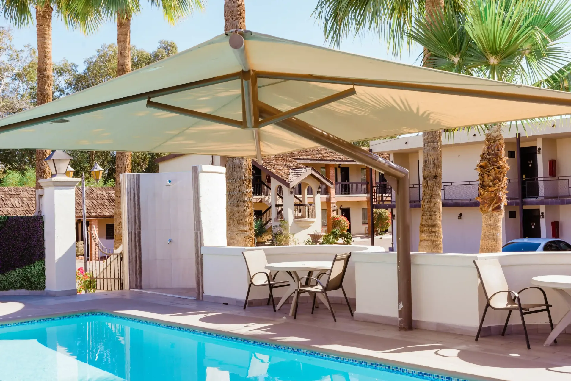 Complejo turístico, una sombrilla voladiza beige  da sombra a un patio junto a la piscina con sillas, mesas y palmeras.