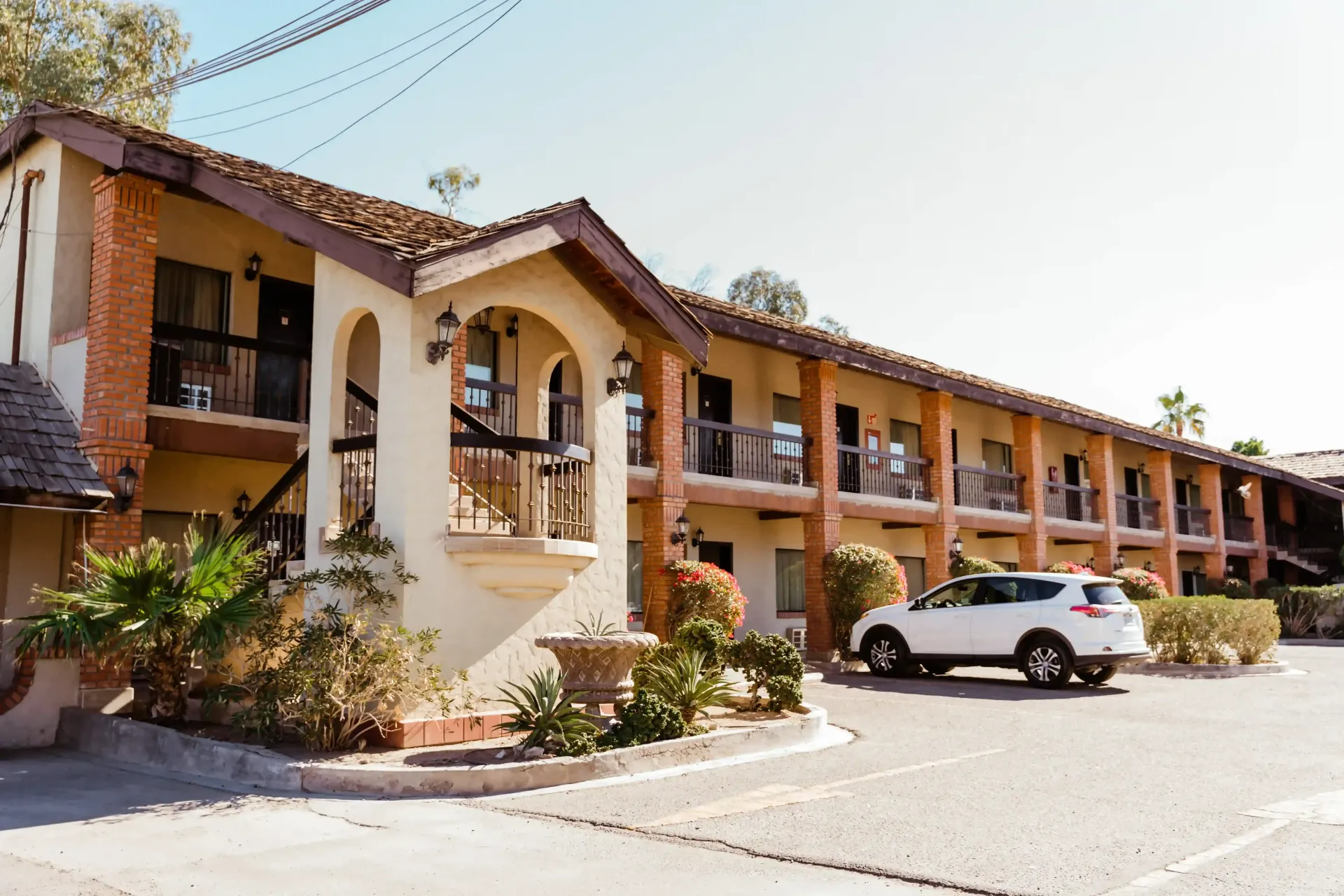 Hotel de dos pisos con fachada de estuco, tejado  marrón y entrada arqueada, con un coche blanco aparcado delante.