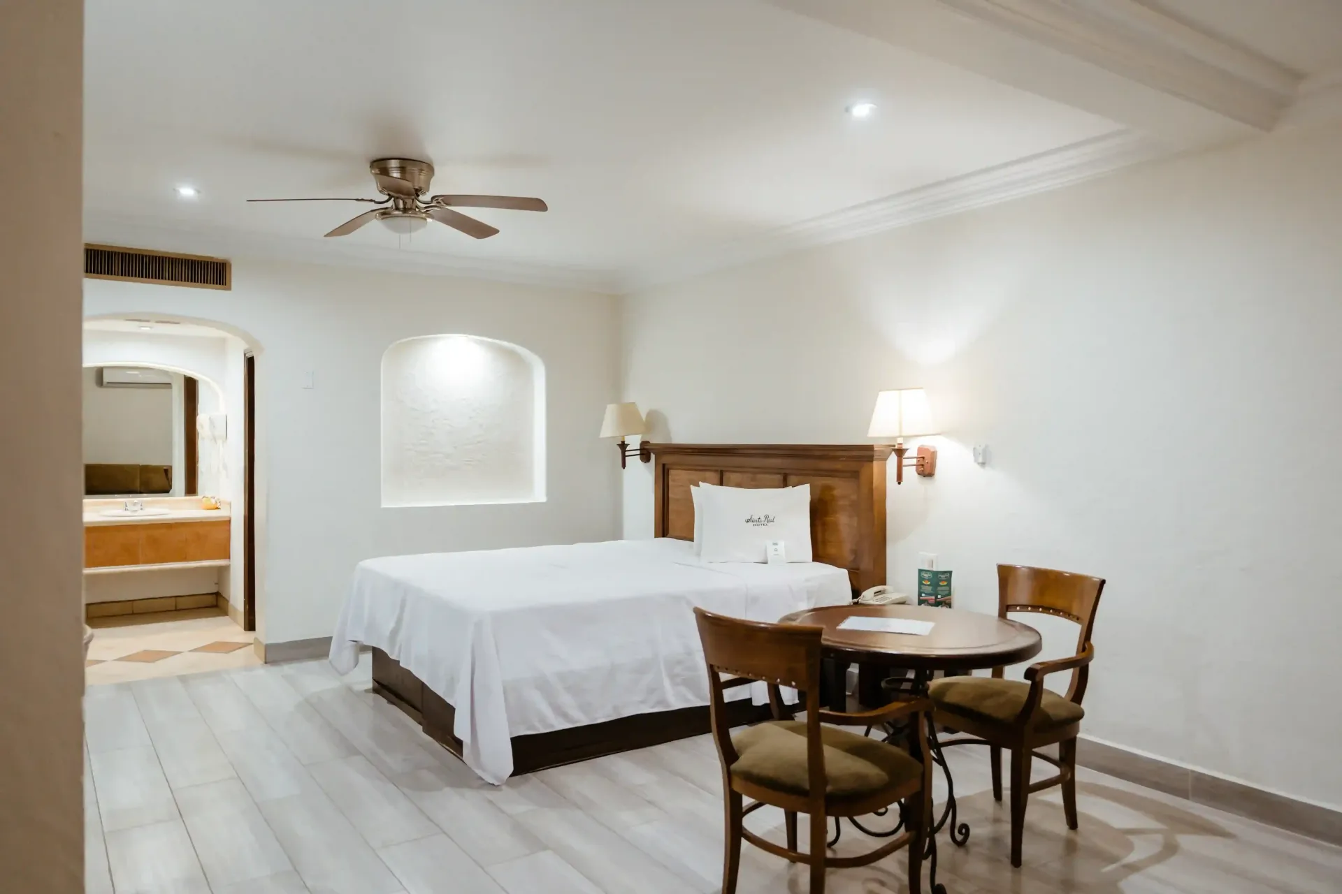 Habitación de hotel con una cama de matrimonio con sábanas blancas, una pequeña mesa de madera con dos sillas 