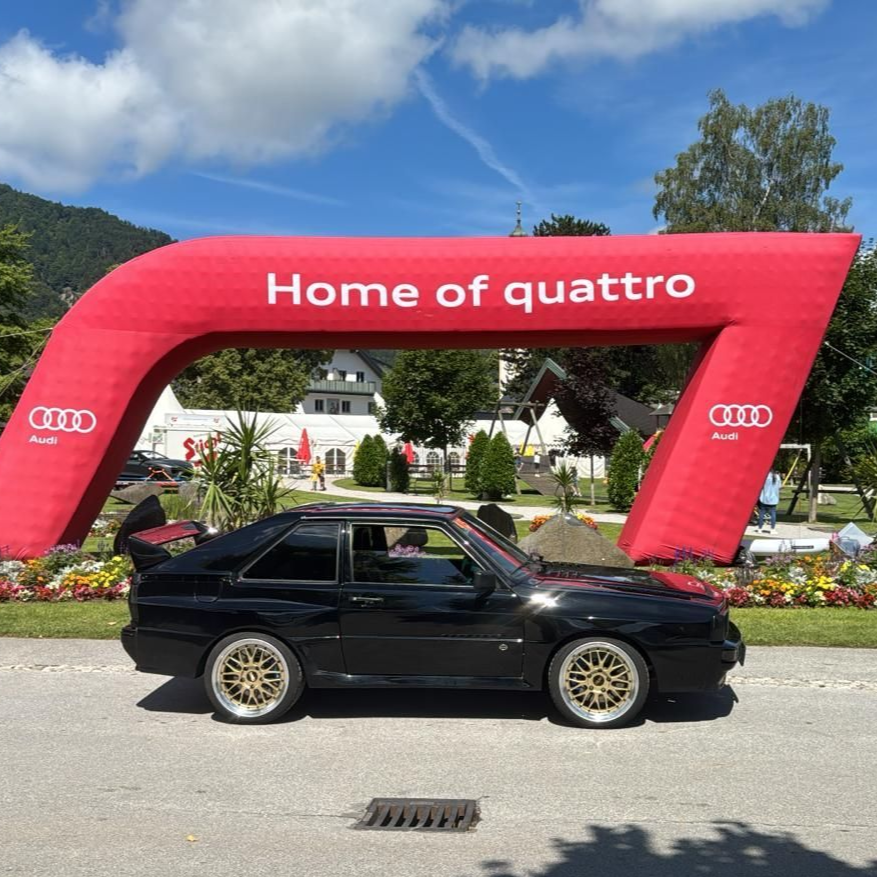 Ein schwarzer Audi Quattro parkt unter einem roten Bogen mit der Aufschrift „Home of quattro“ vor einem klaren blauen Himmel.