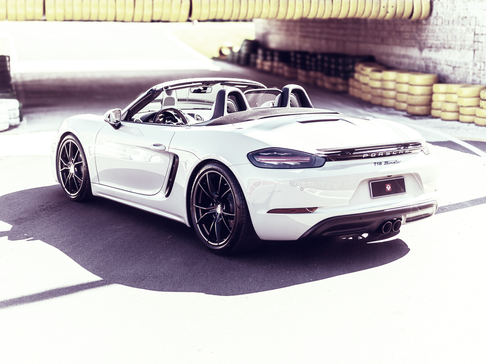 Ein hellgraues Porsche 718 Boxster Cabriolet parkt auf einer Rennstrecke; im Hintergrund sind gelbe Leitplanken zu sehen.
