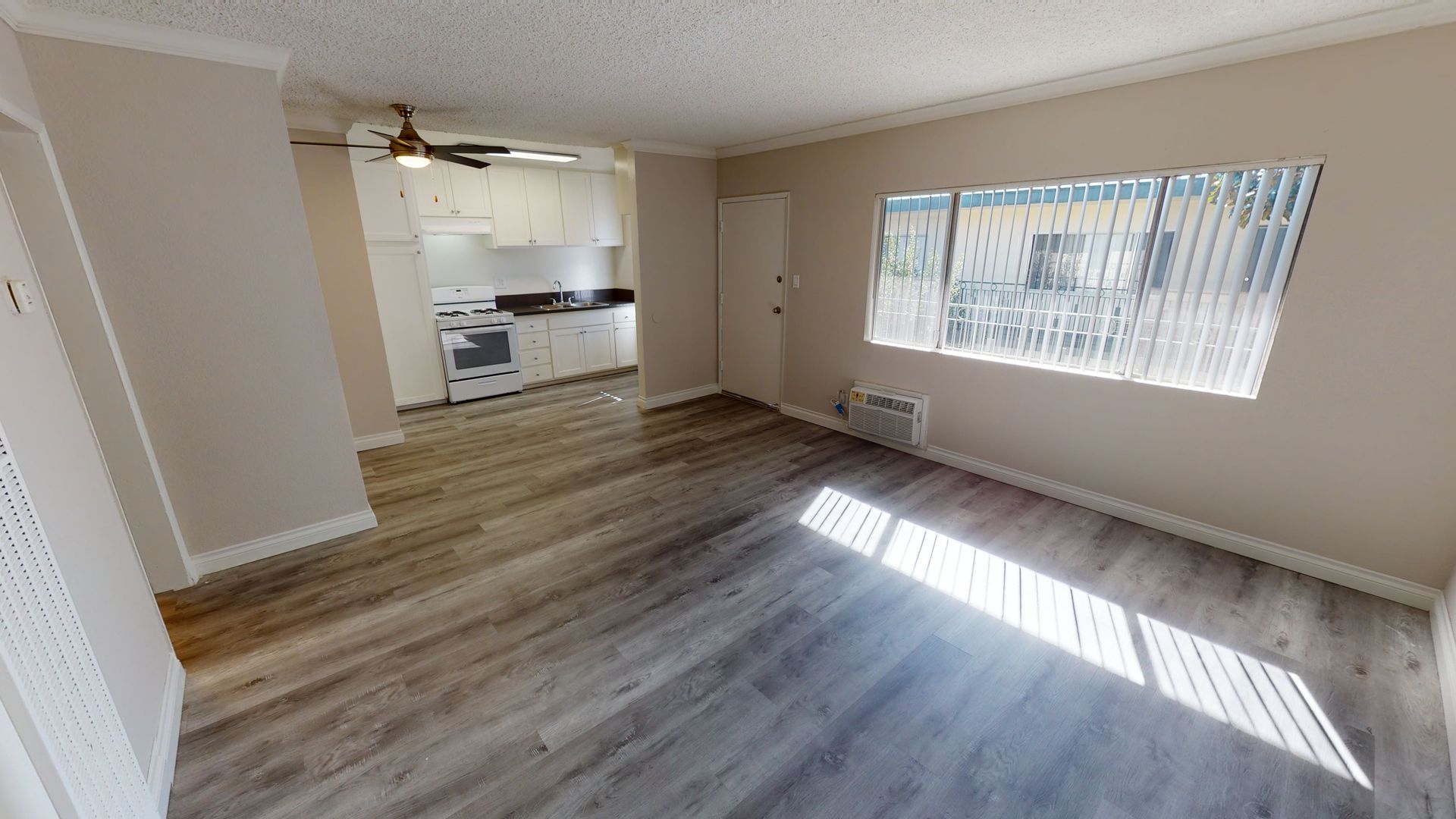 Living room with wooden floors, kitchen in the background, window with blinds, and a door.