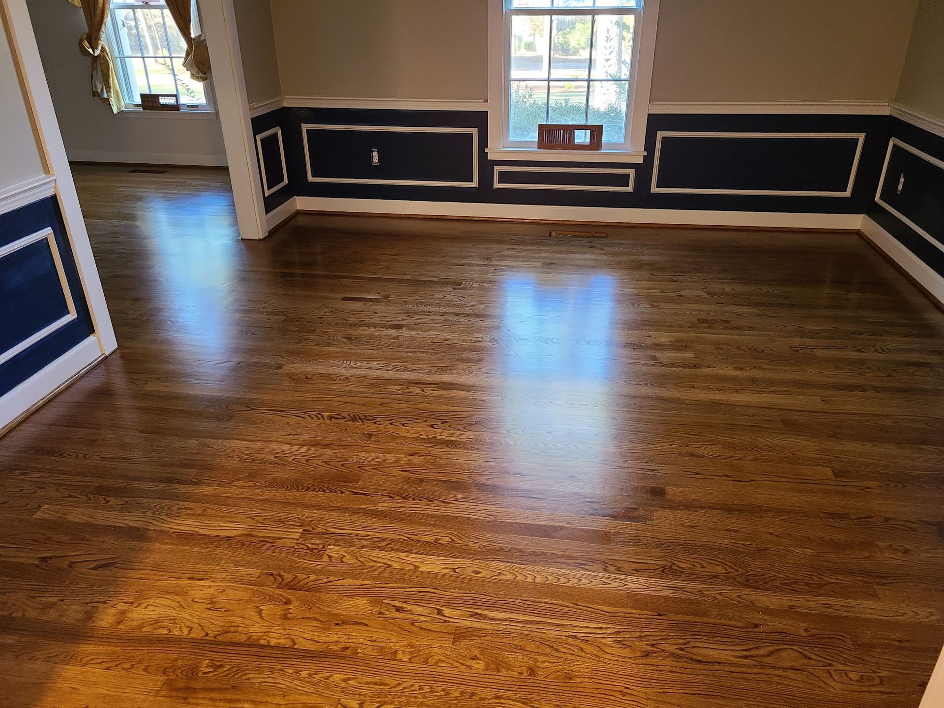 Refinished hardwood floors reflect light in a dining room with dark blue wainscoting and white trim.