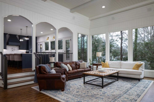 Sunroom with leather sofa, white couch, large windows overlooking trees, arched doorways leading to kitchen.