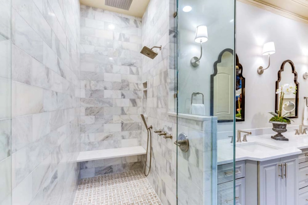 Marble-tiled shower with a bench, glass door, and hand shower. Adjacent to a vanity with mirrors and sconces.
