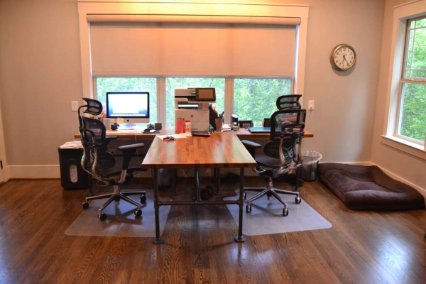 Home office with two desks, chairs, and monitors near a window. Wooden floors, a dog bed.