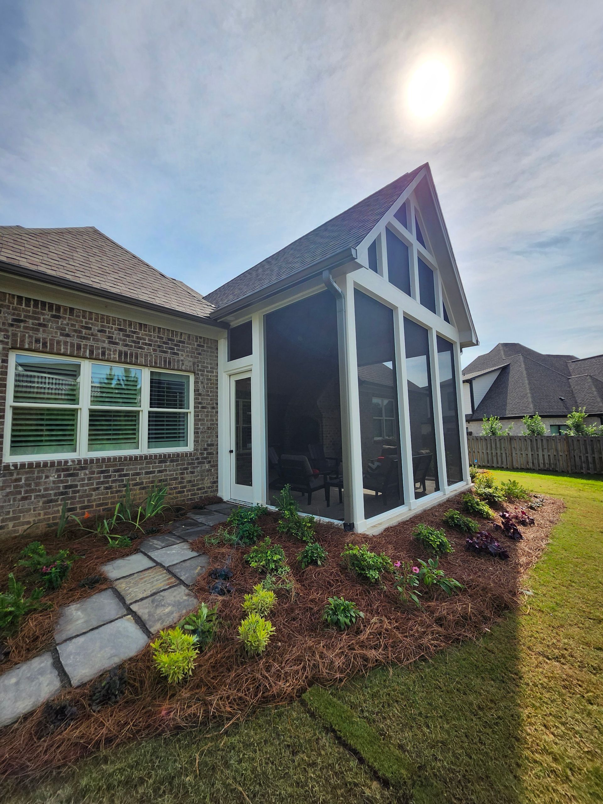 Screened-in porch attached to a brick house with flowerbeds and sunny sky.