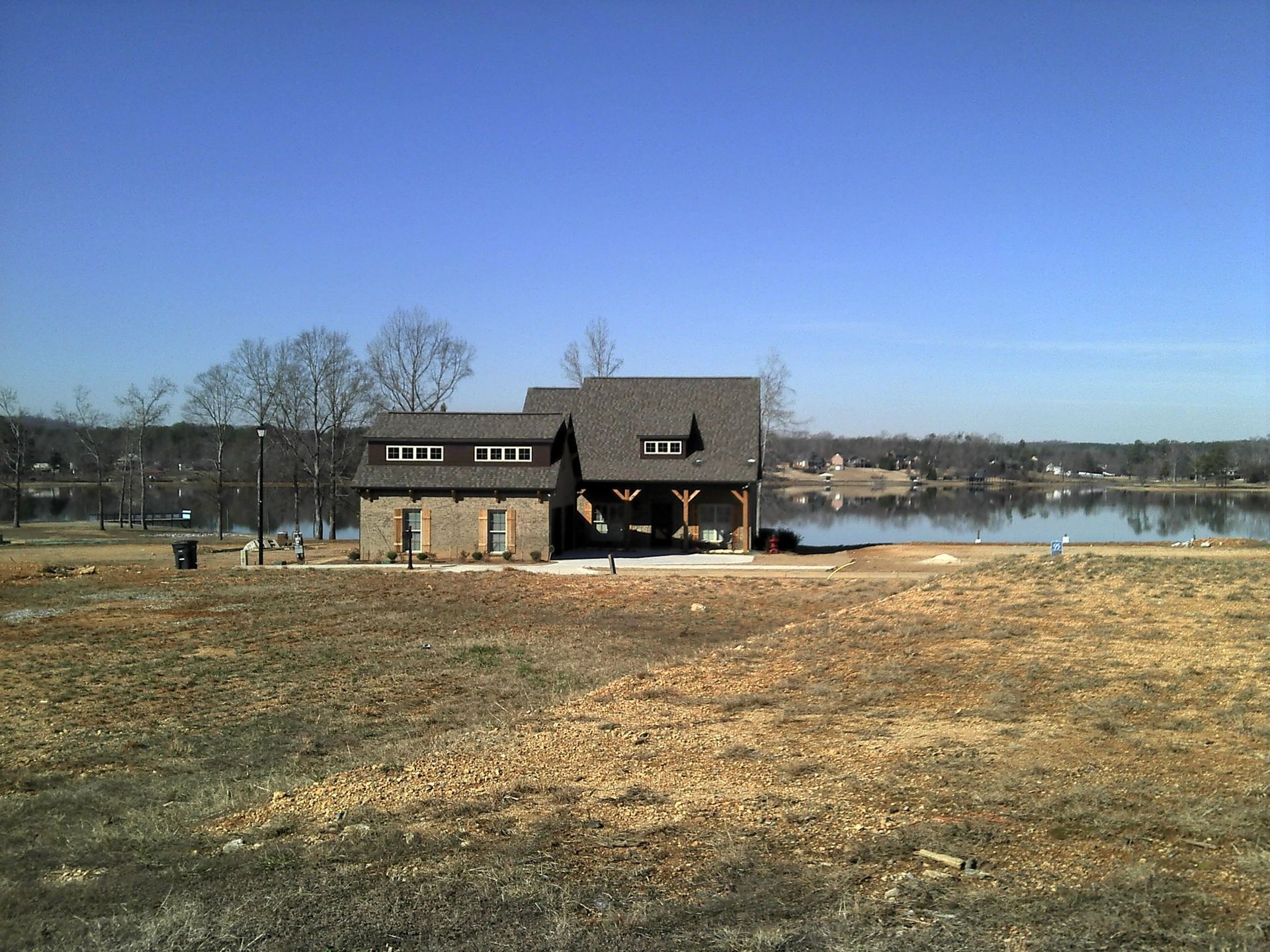 Lakeside building with stone facade, under a gray roof, set against a blue sky. Bare trees line the shore.