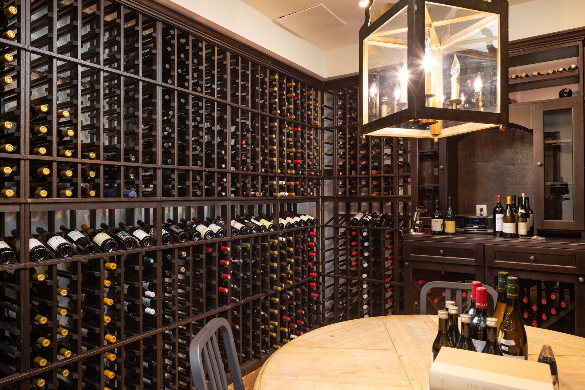 Wine cellar with rows of bottles on metal racks, a round table, and a decorative light fixture.