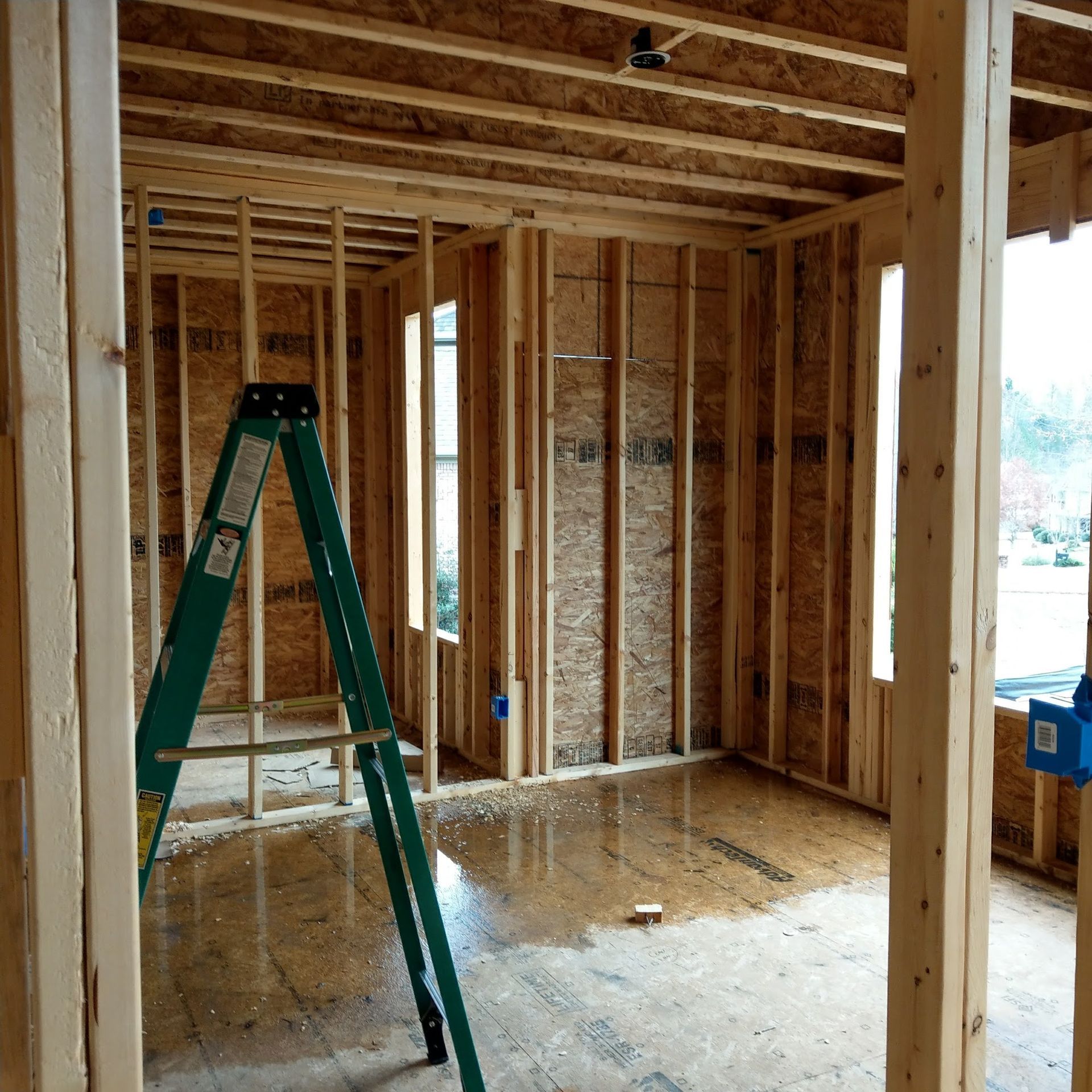 Interior framing of a room under construction with a green ladder and water on the floor.