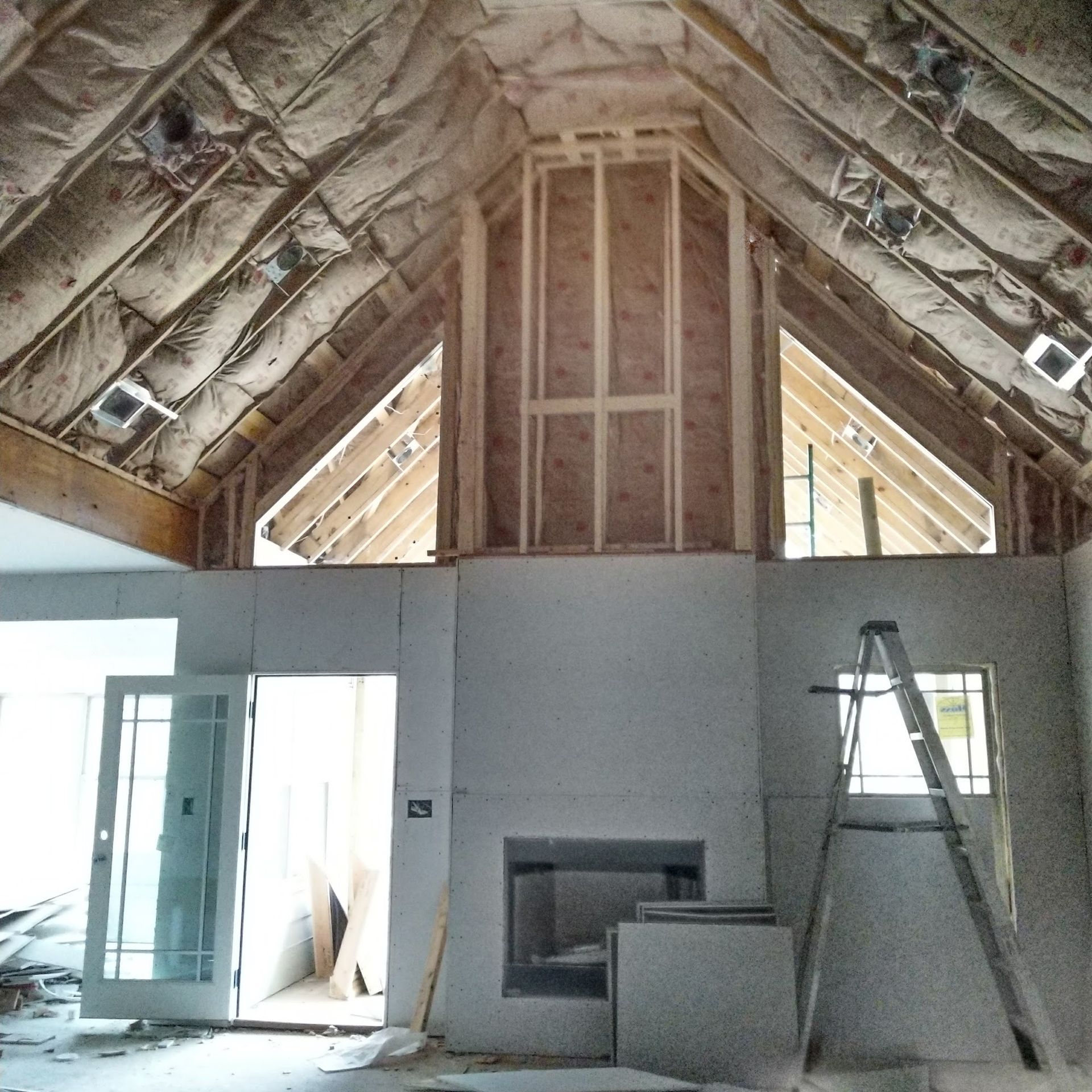 Interior of a house under construction; exposed framing, insulation, fireplace, ladder, and a door are visible.