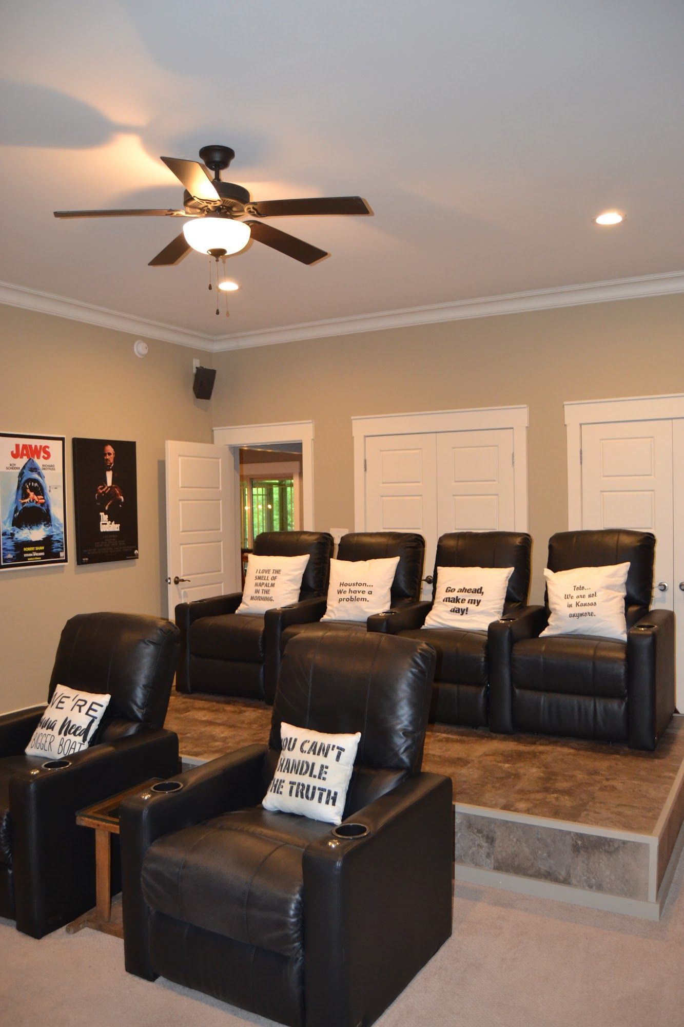 Home theater with black recliners on a tiered platform. Movie posters, tan walls, and a ceiling fan are visible.
