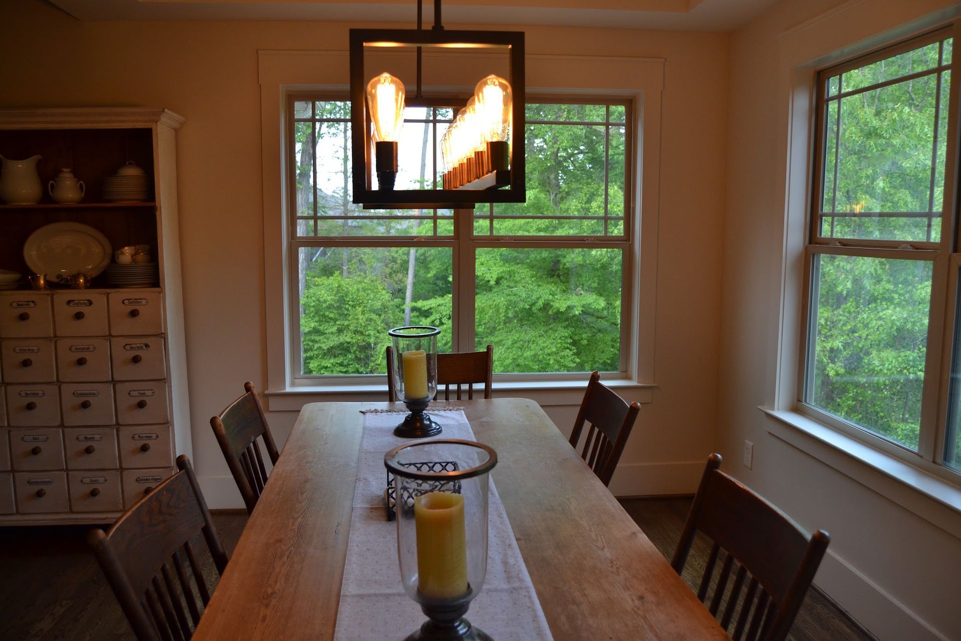 Dining room with wooden table, chairs, and decorative candles, beneath a modern light fixture and windows