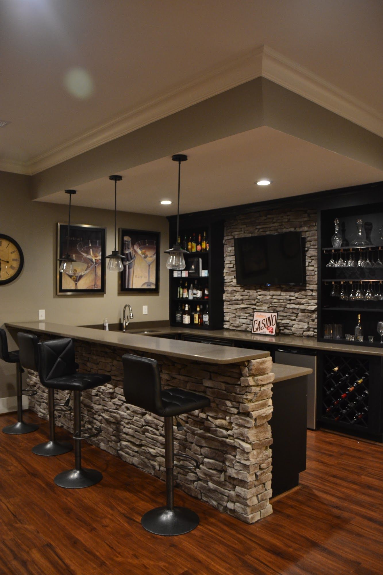 Basement bar with stone facade, seating, shelves, and a TV. Features dark wood floors and bar stools.