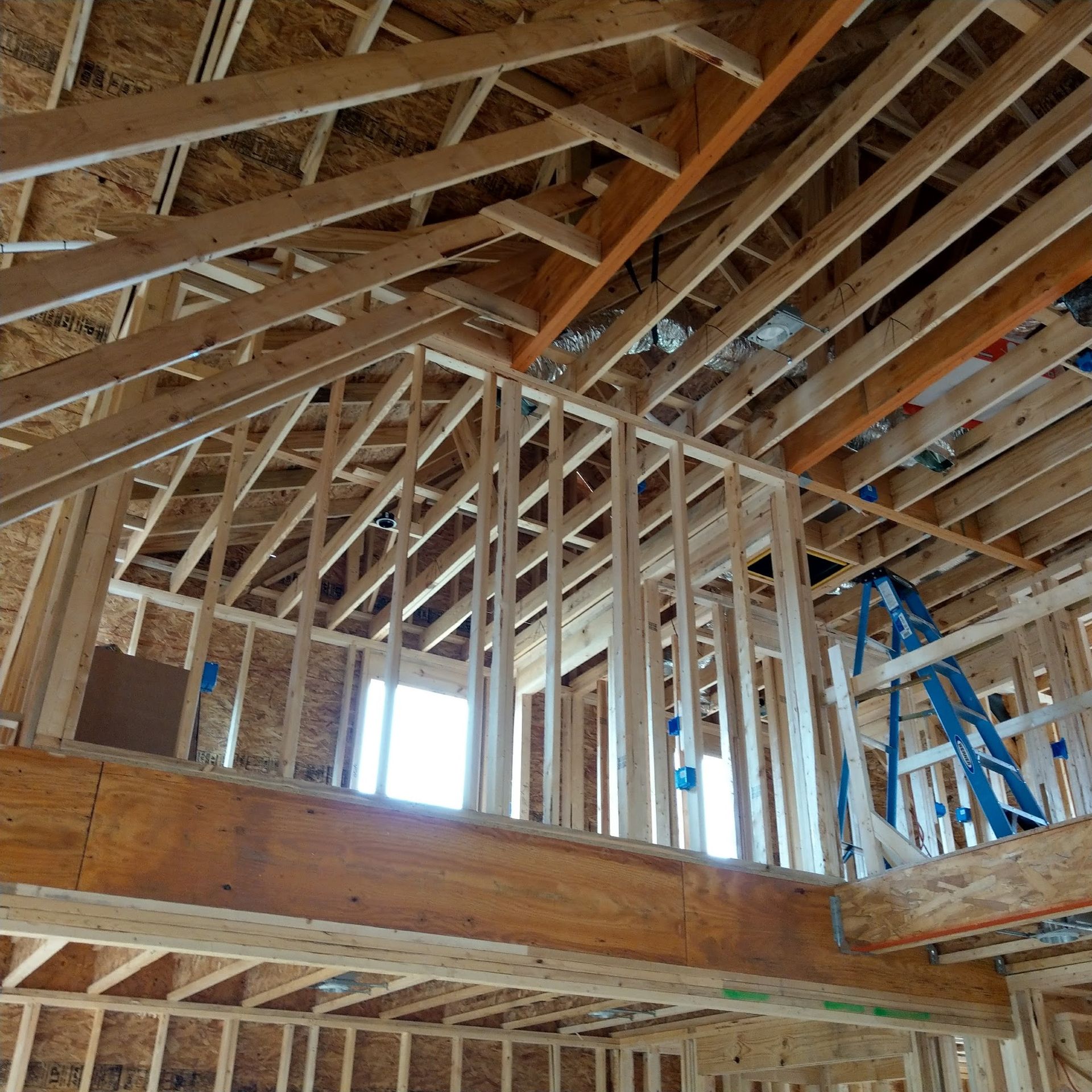 Wooden framing of a house under construction; interior view. Studs, rafters, and beams visible. Blue ladder present.