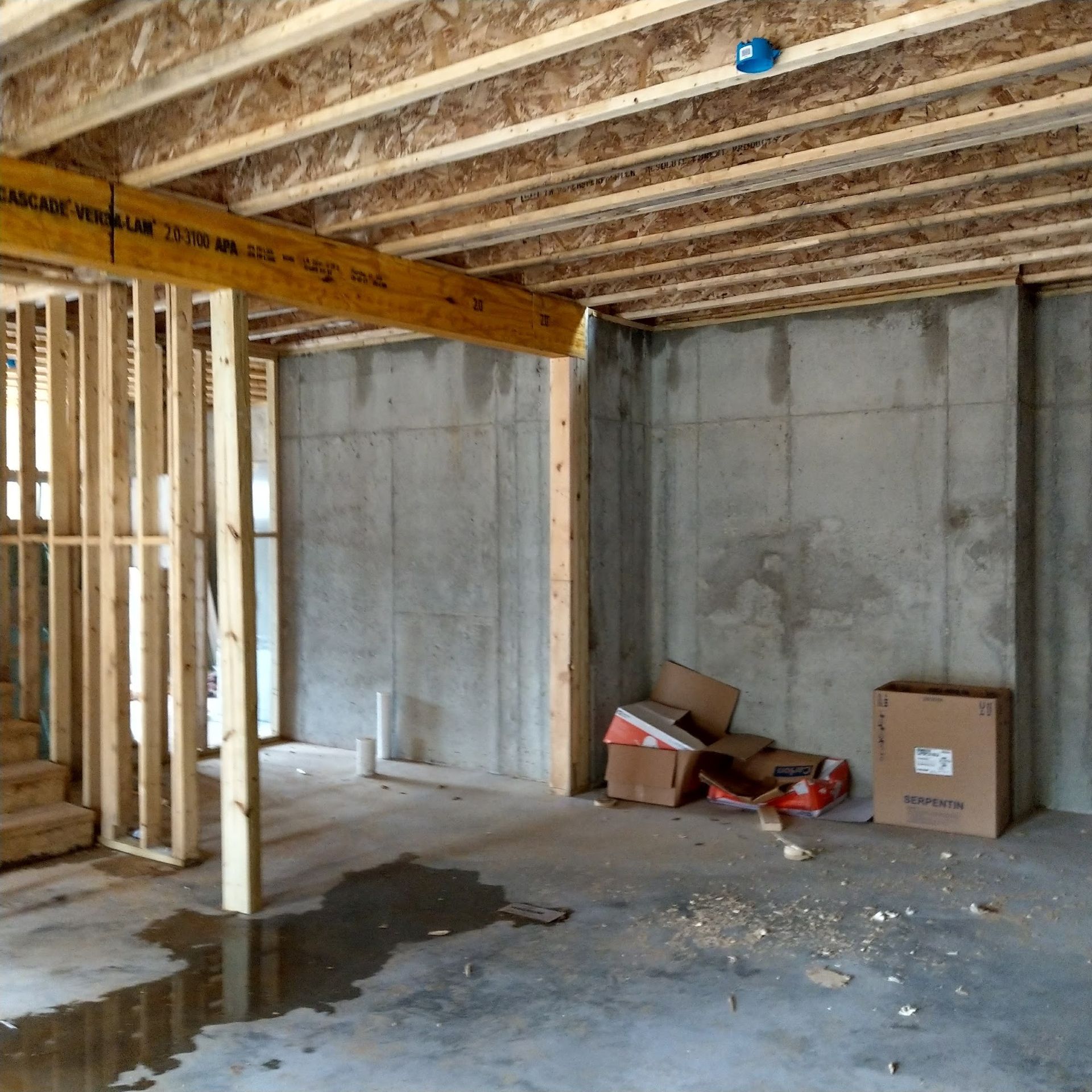 Basement under construction, concrete walls and floor. Exposed wood beams and framing, boxes on floor.