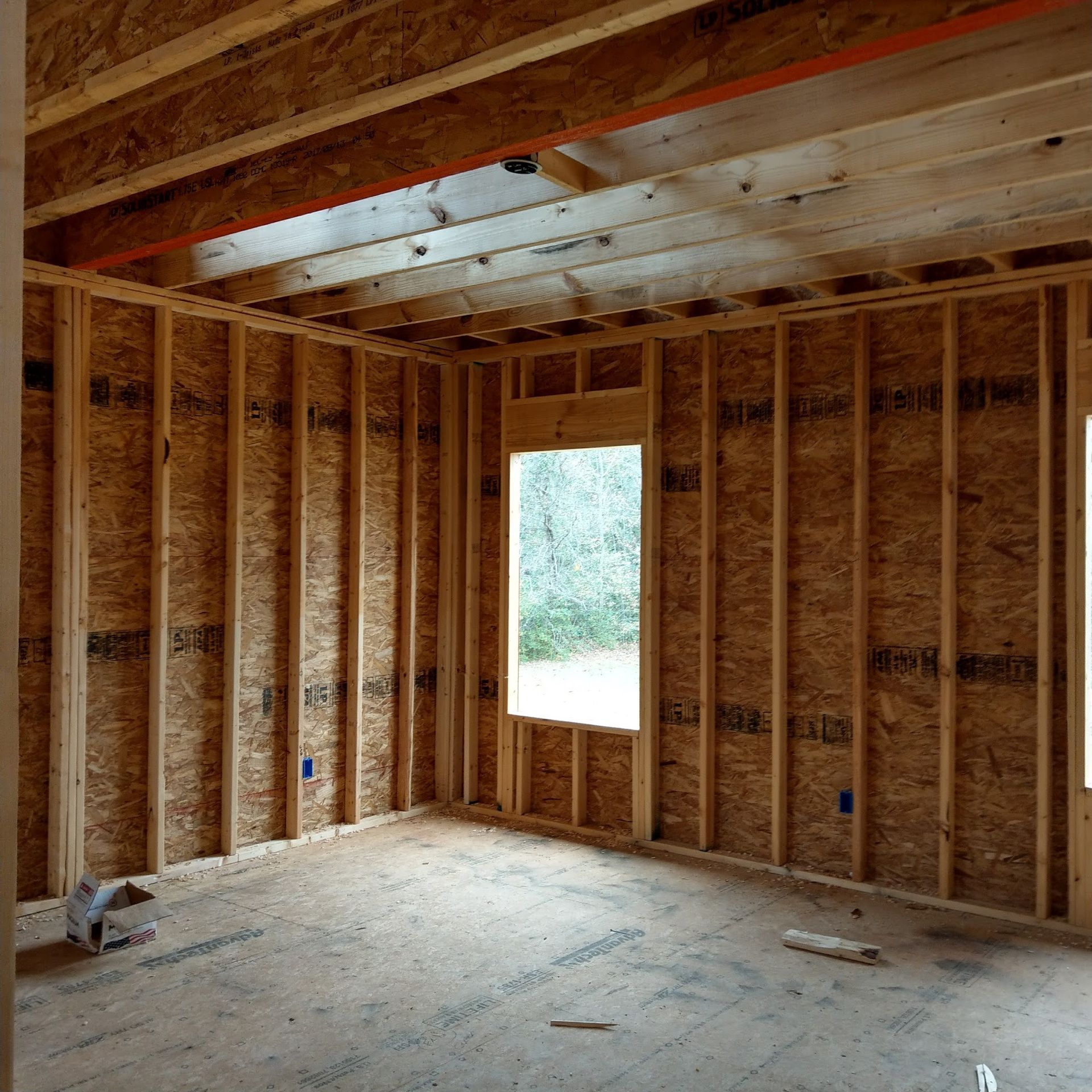 Interior of a room under construction, showing wooden studs and unfinished walls and ceiling. A window is in the center.