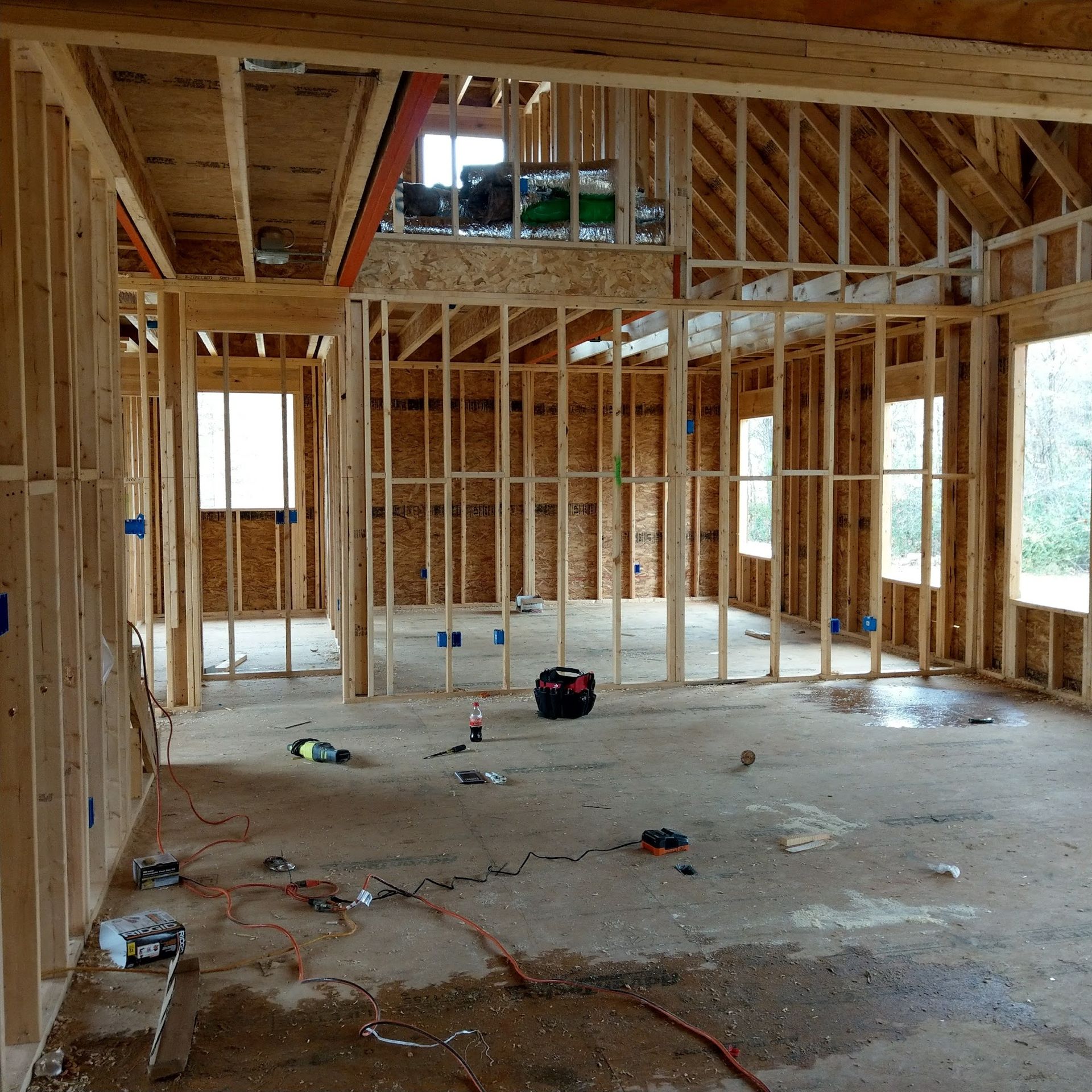 Interior view of a house under construction. Wooden framing, bare concrete floor, and tools scattered.