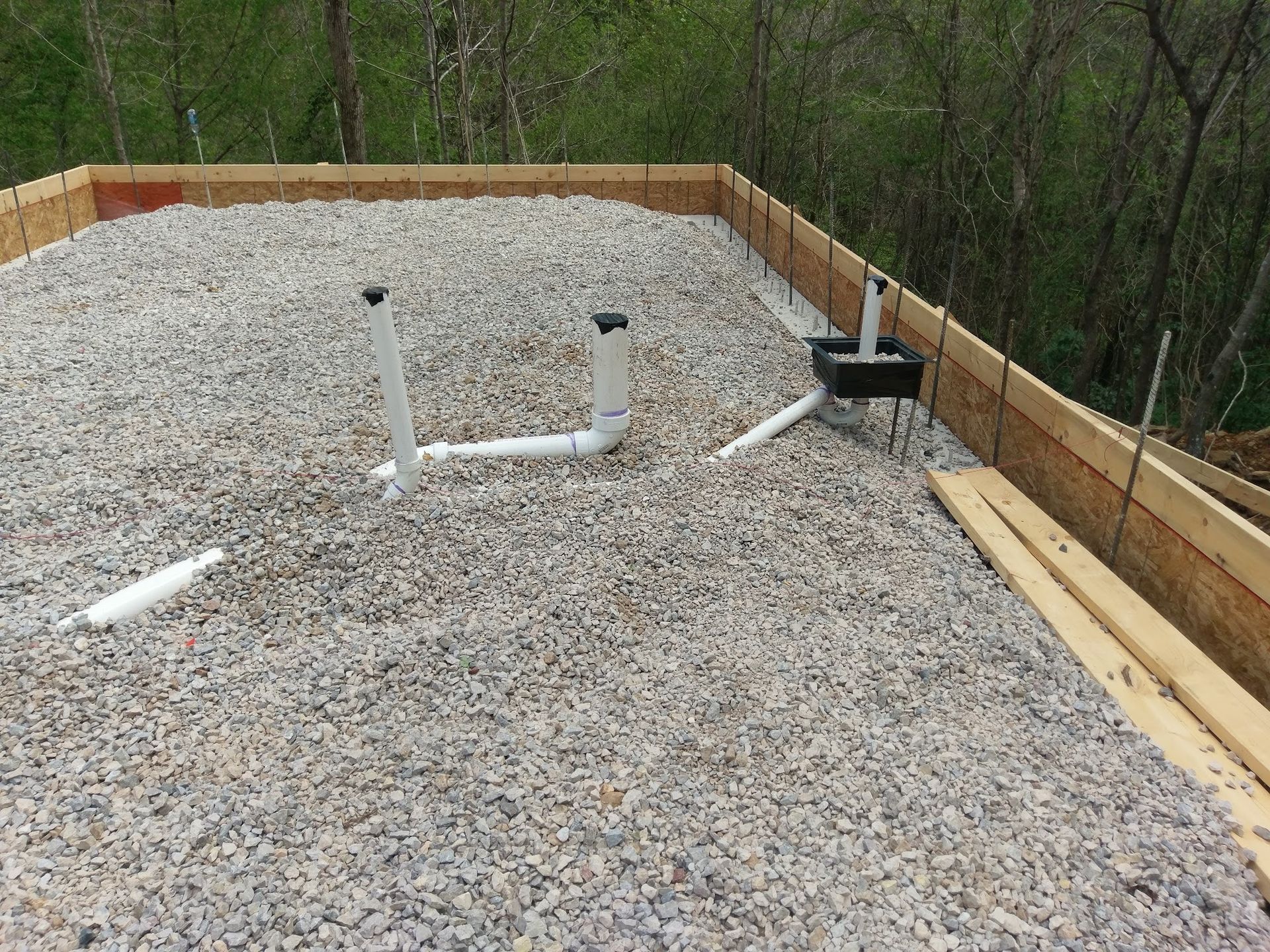 Gravel-filled concrete form with white PVC pipes for plumbing, surrounded by wooden framing in an outdoor setting.