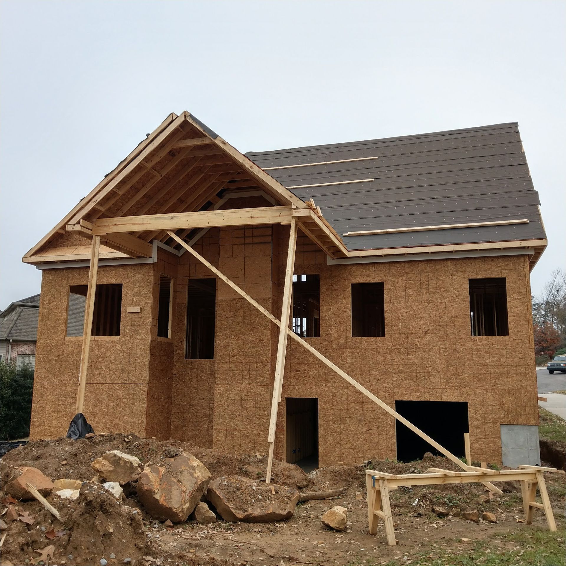 Two-story house under construction; exposed wood frame, dark brown roof, windows, and front porch supports.