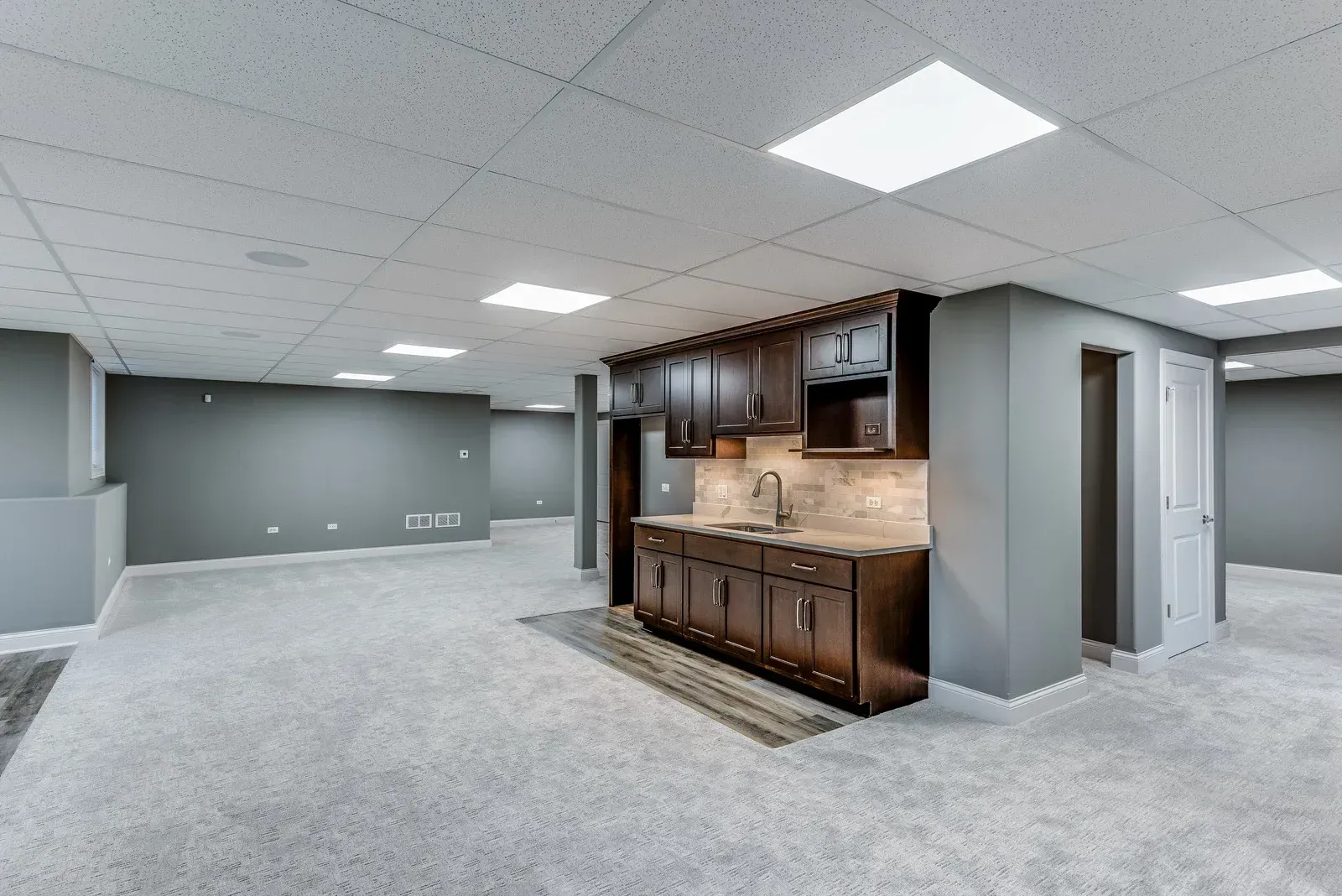 Spacious finished basement with a bar, gray walls, and carpet. Recessed lighting and a small doorway are visible.