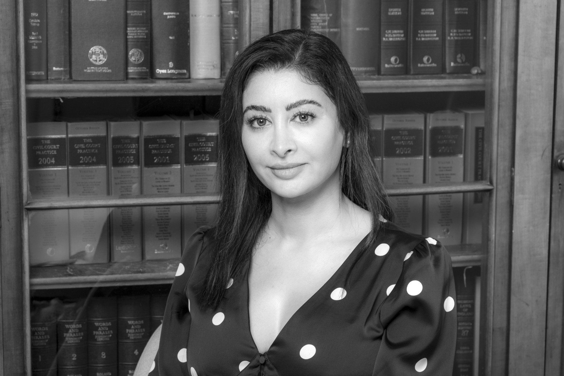 Woman in polka-dot blouse seated before a bookshelf, looking at the camera.