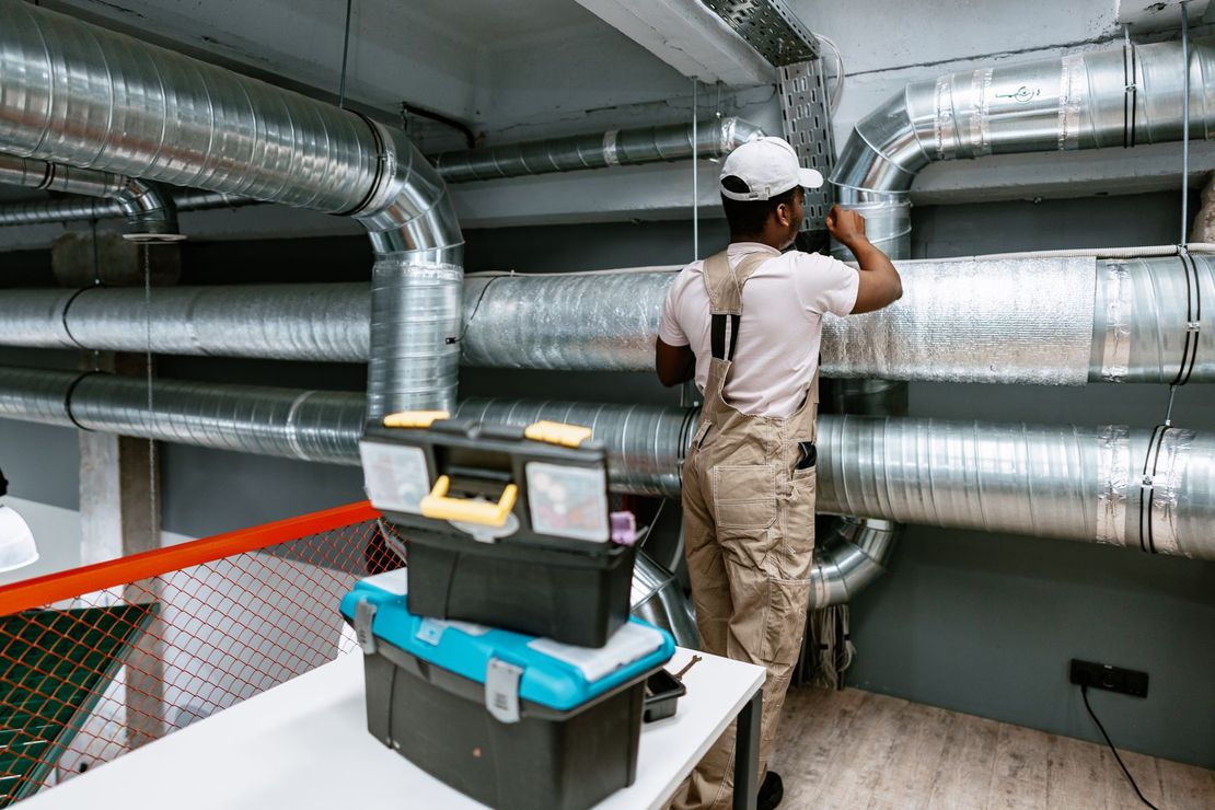 Technician inspecting large silver HVAC ducts in a mechanical room with tools on a cart