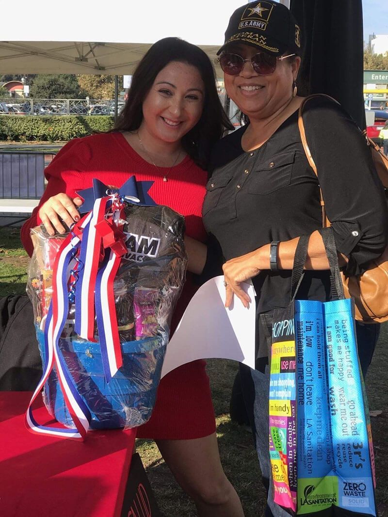 Two women smile, one holding a decorated backpack, the other holding a list, outdoors.
