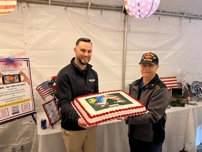 Two men holding a cake with a military image, indoors with patriotic decorations.