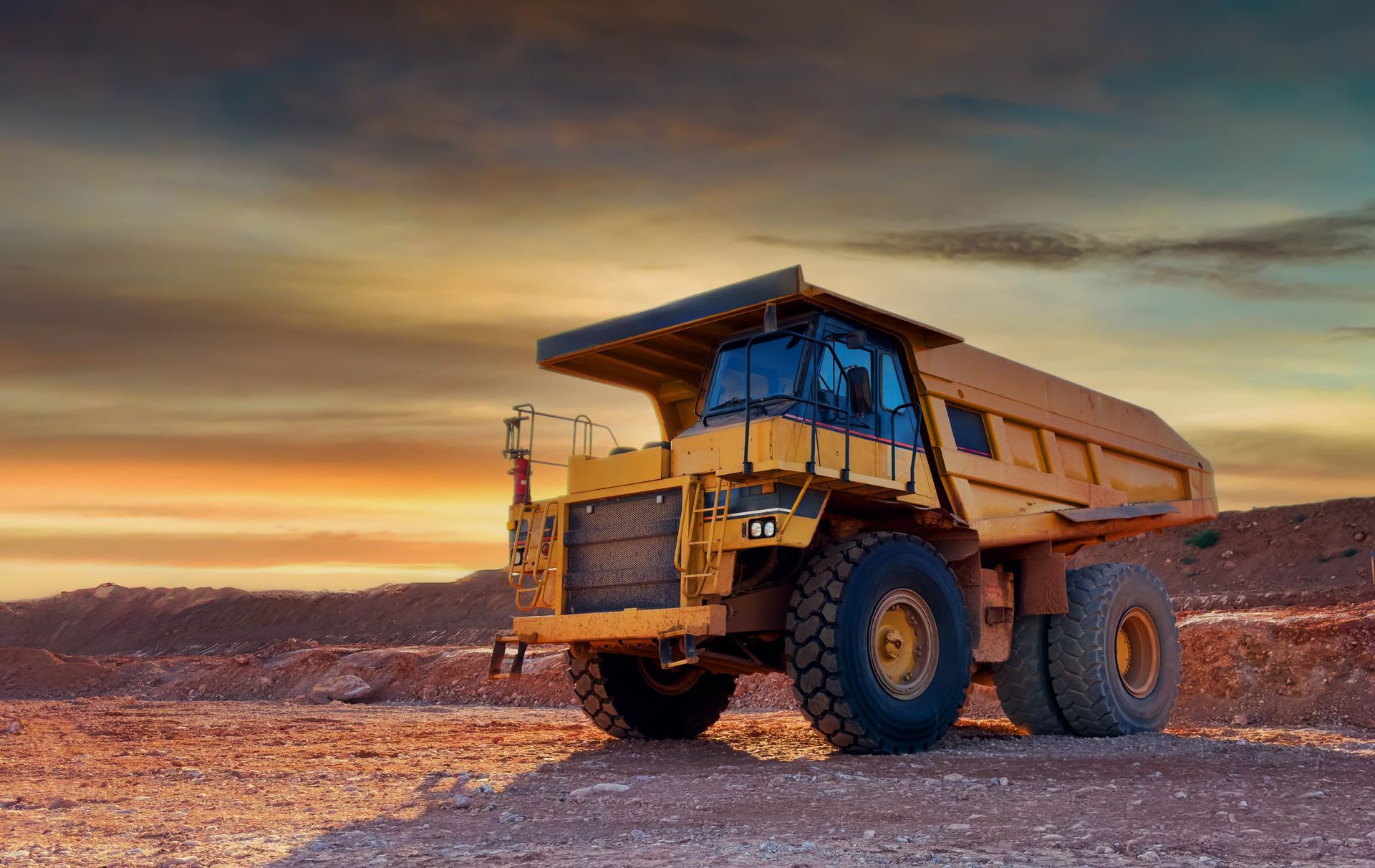 Large yellow dump truck on a rocky site at sunset, with dramatic sky and mountains in the background