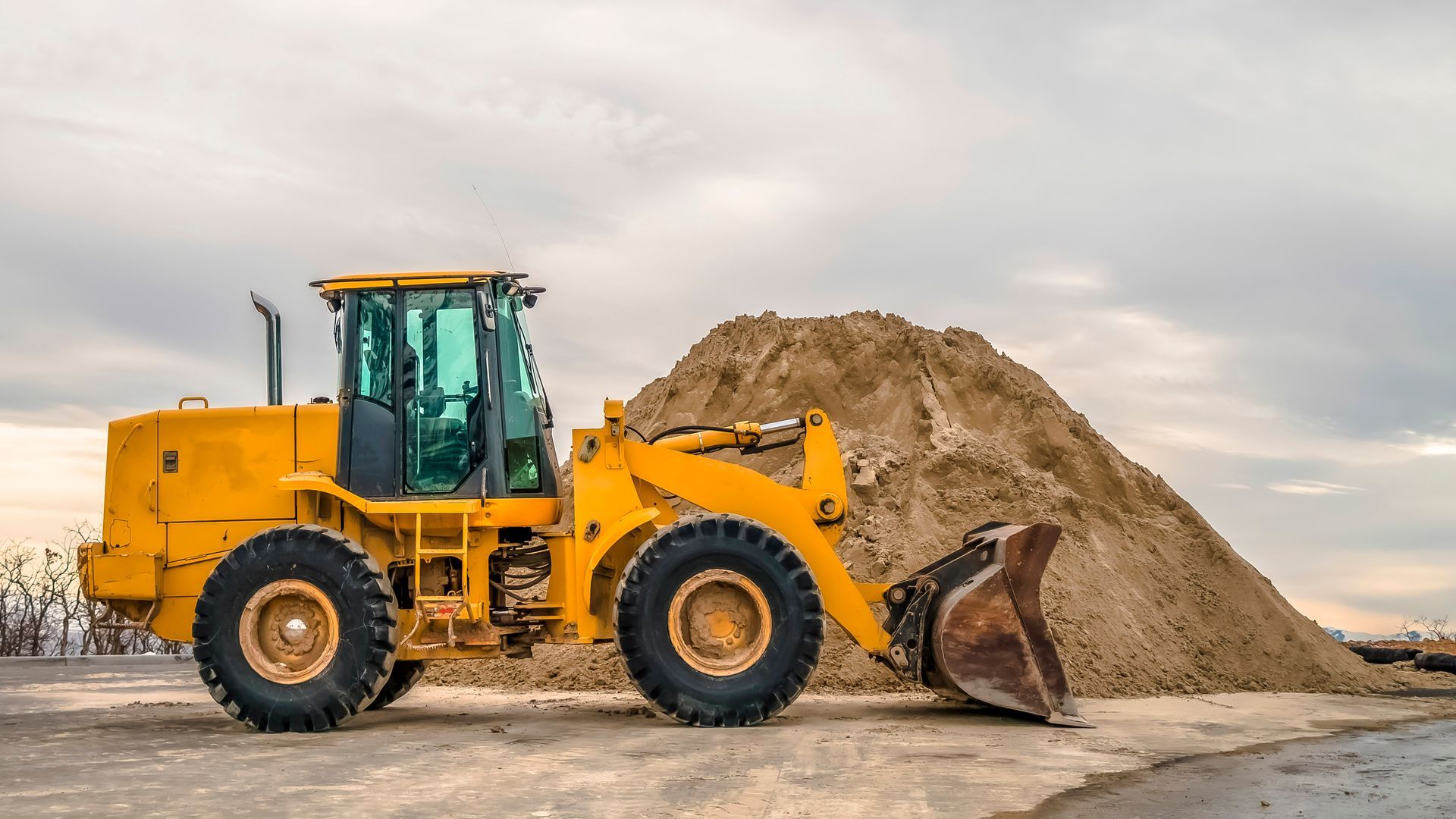 Yellow front loader parked beside a large sand pile under an overcast sky