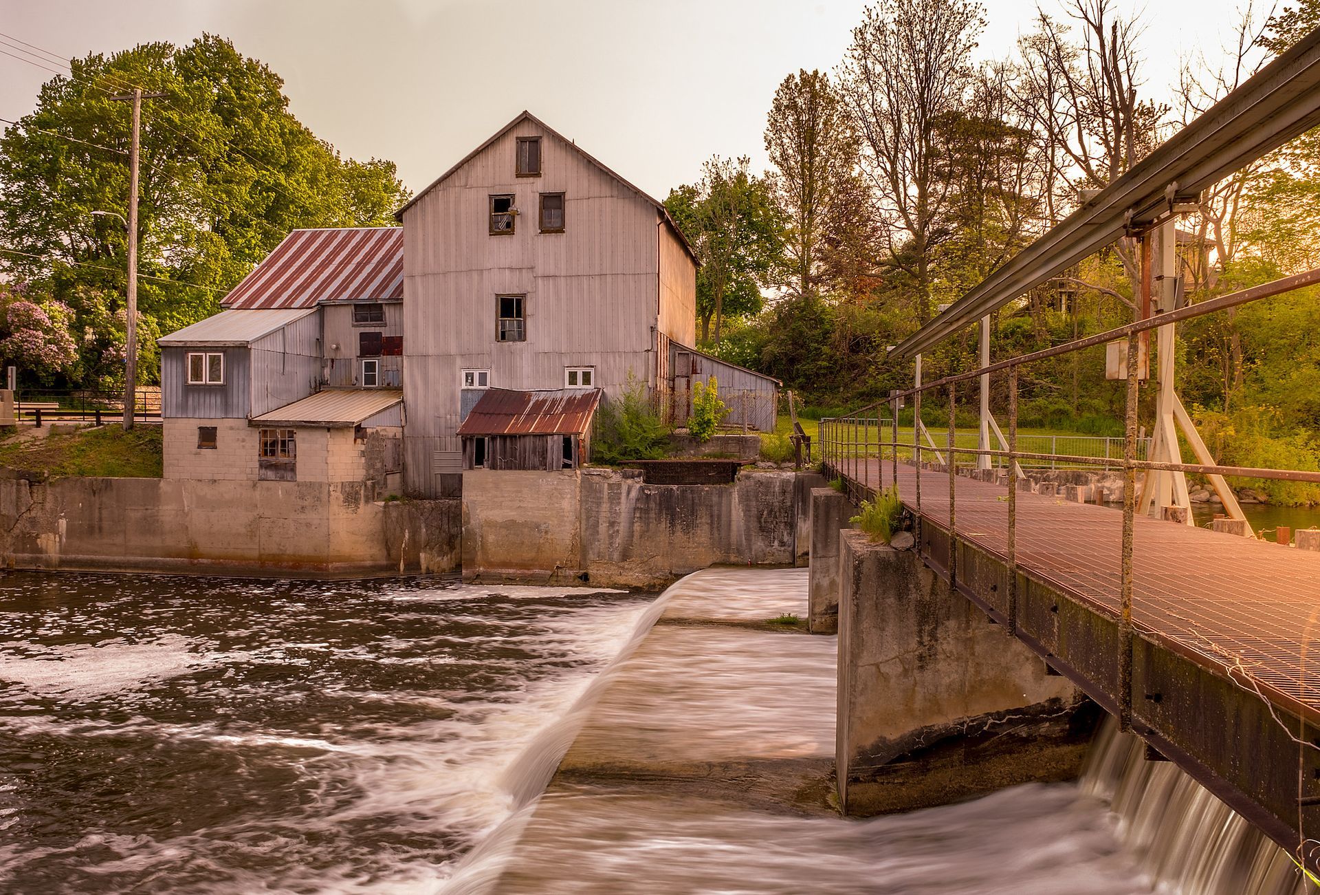 Old mill by a rushing river, with a bridge and trees in warm evening light