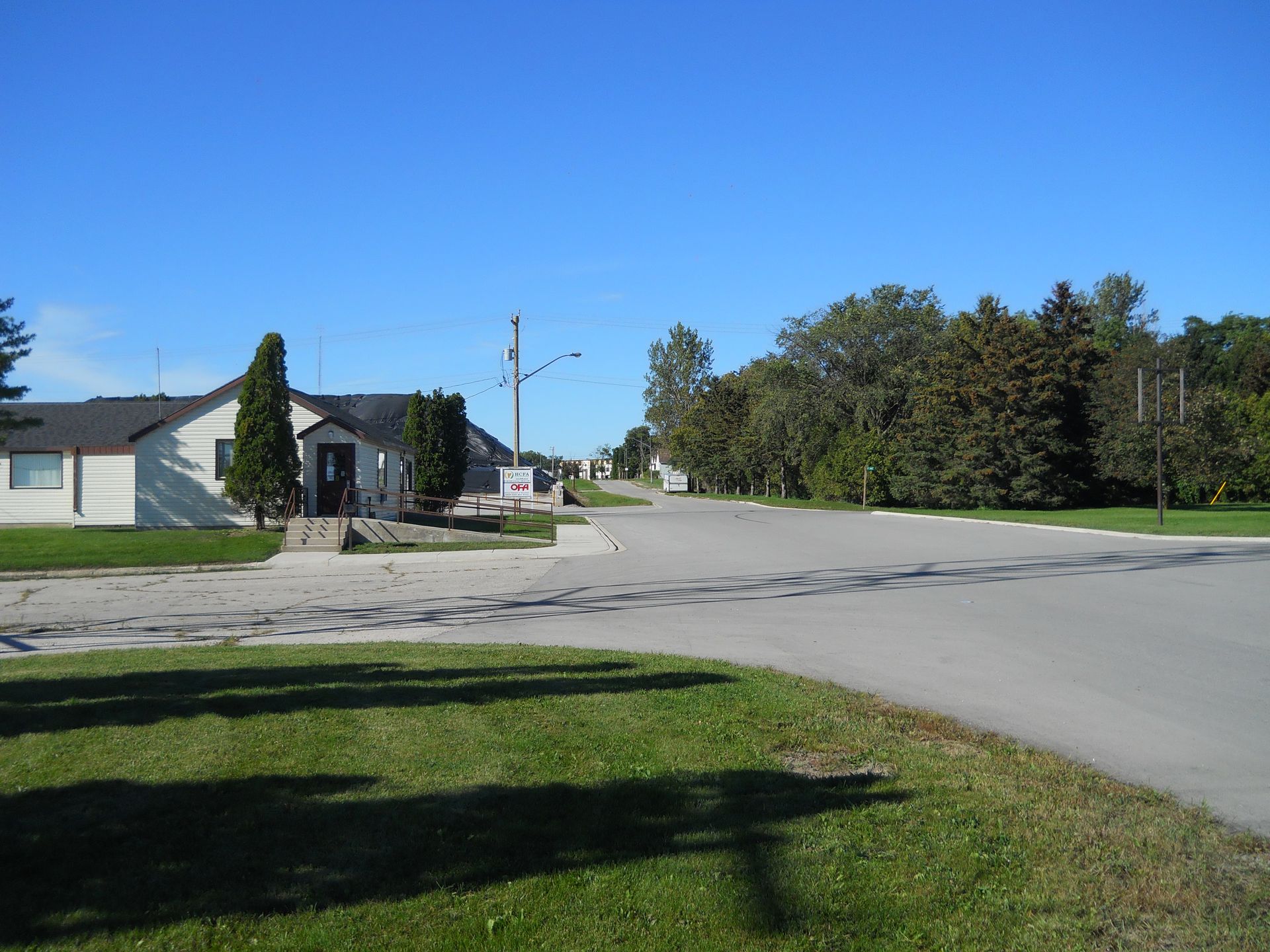 Quiet residential street with a house, trees, and a wide empty road under a clear blue sky