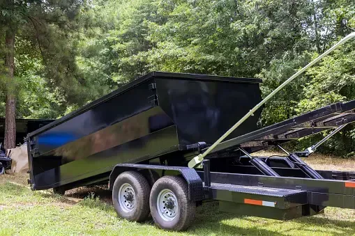 Black dump trailer with raised bed parked on grass in a wooded area