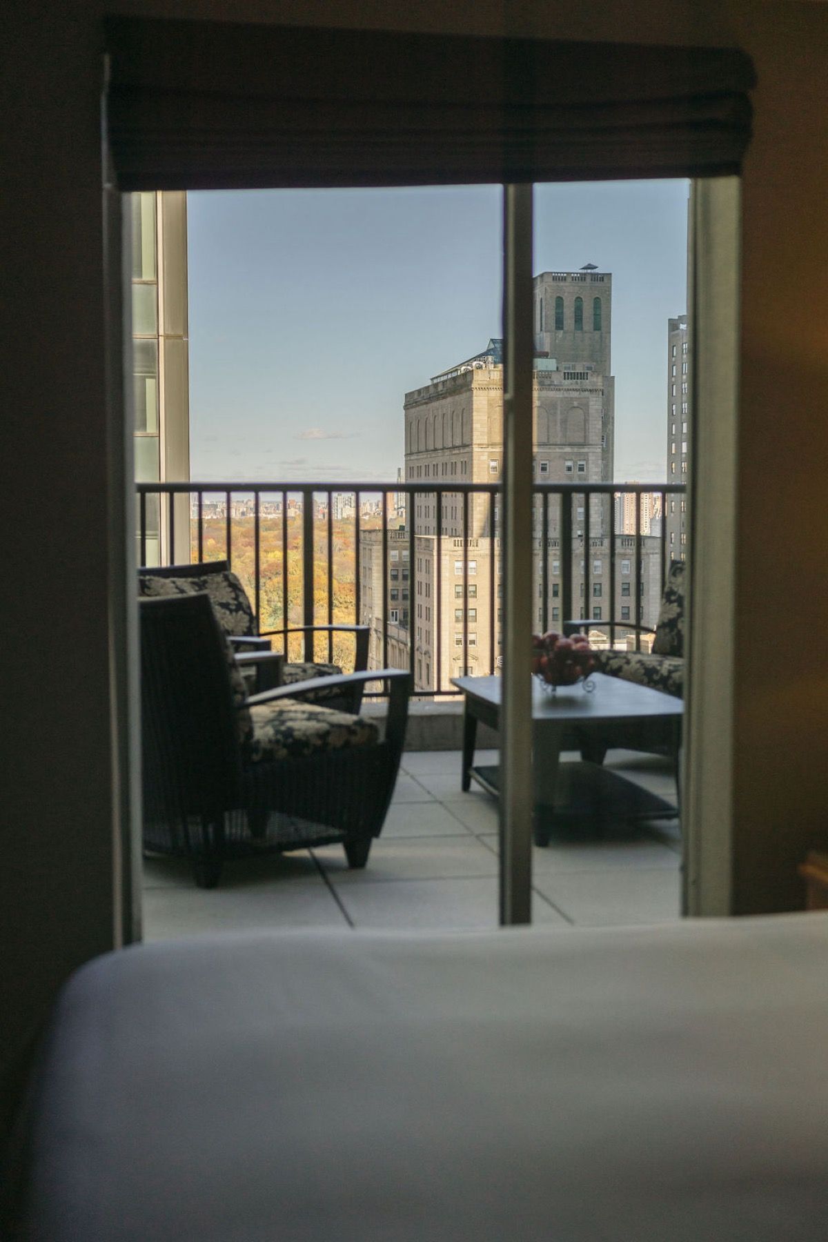 Hotel balcony view through open doors to city skyline at sunset, with wicker chairs and bed in foreground