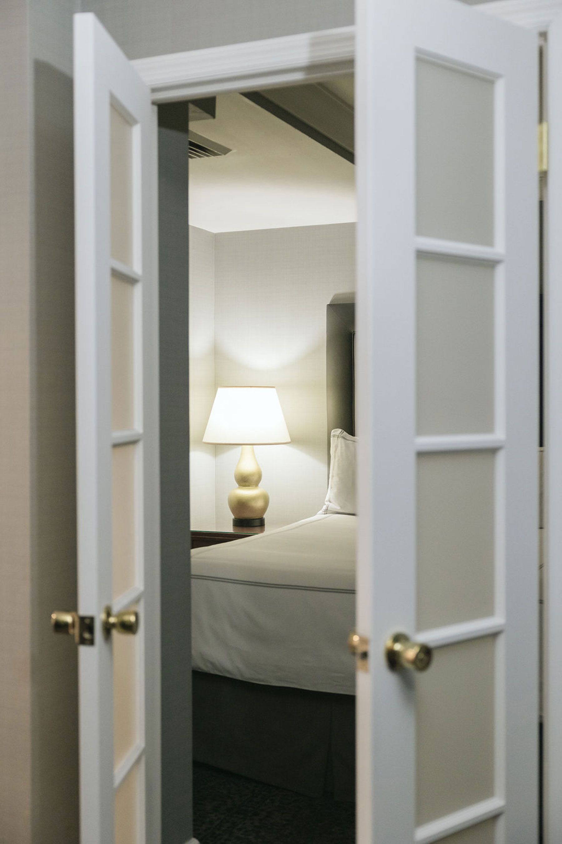 A hotel room with a bed, lamp, and nightstand visible through an open door.