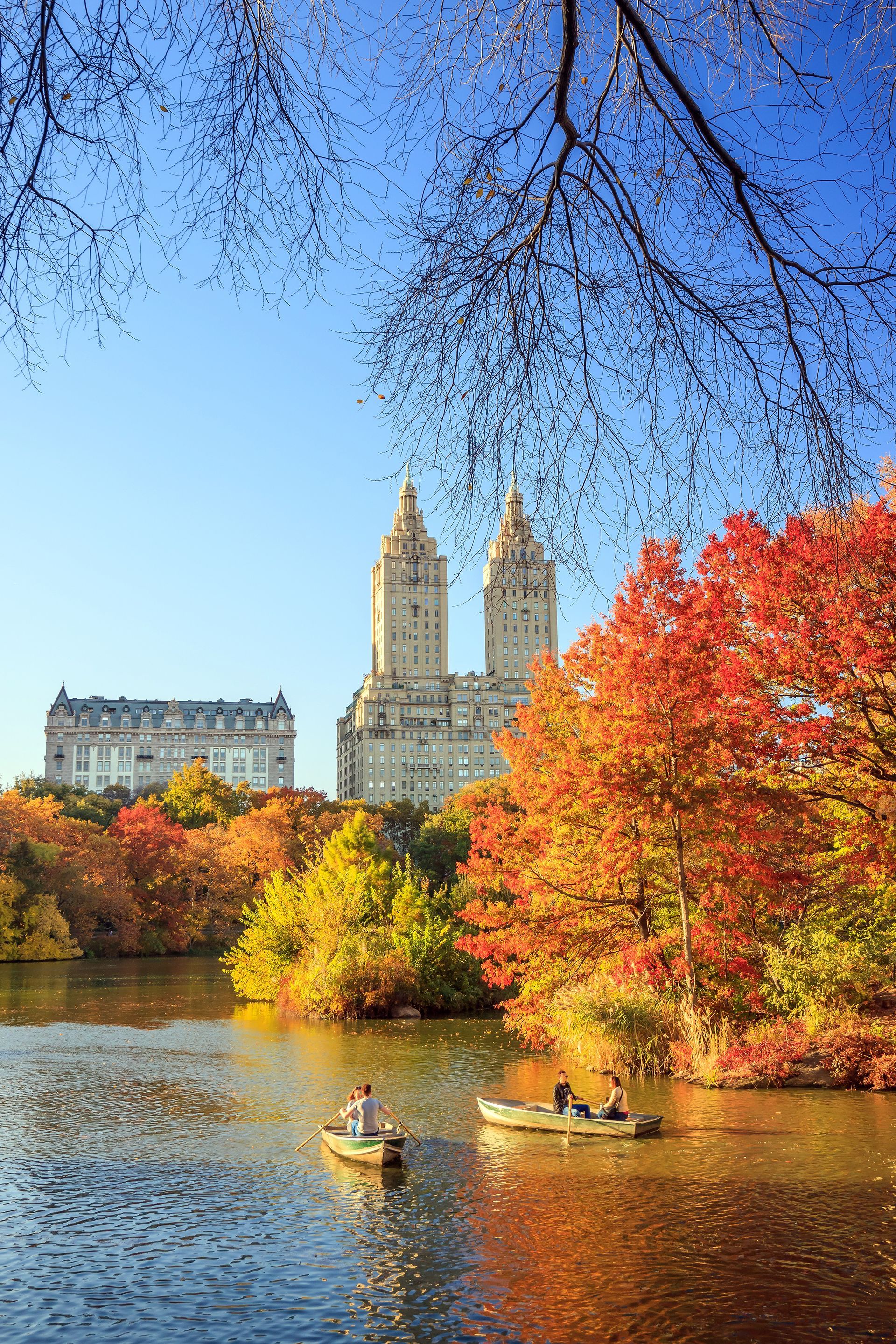 Autumn park lake with kayaks, colorful trees, and twin towers under a clear blue sky