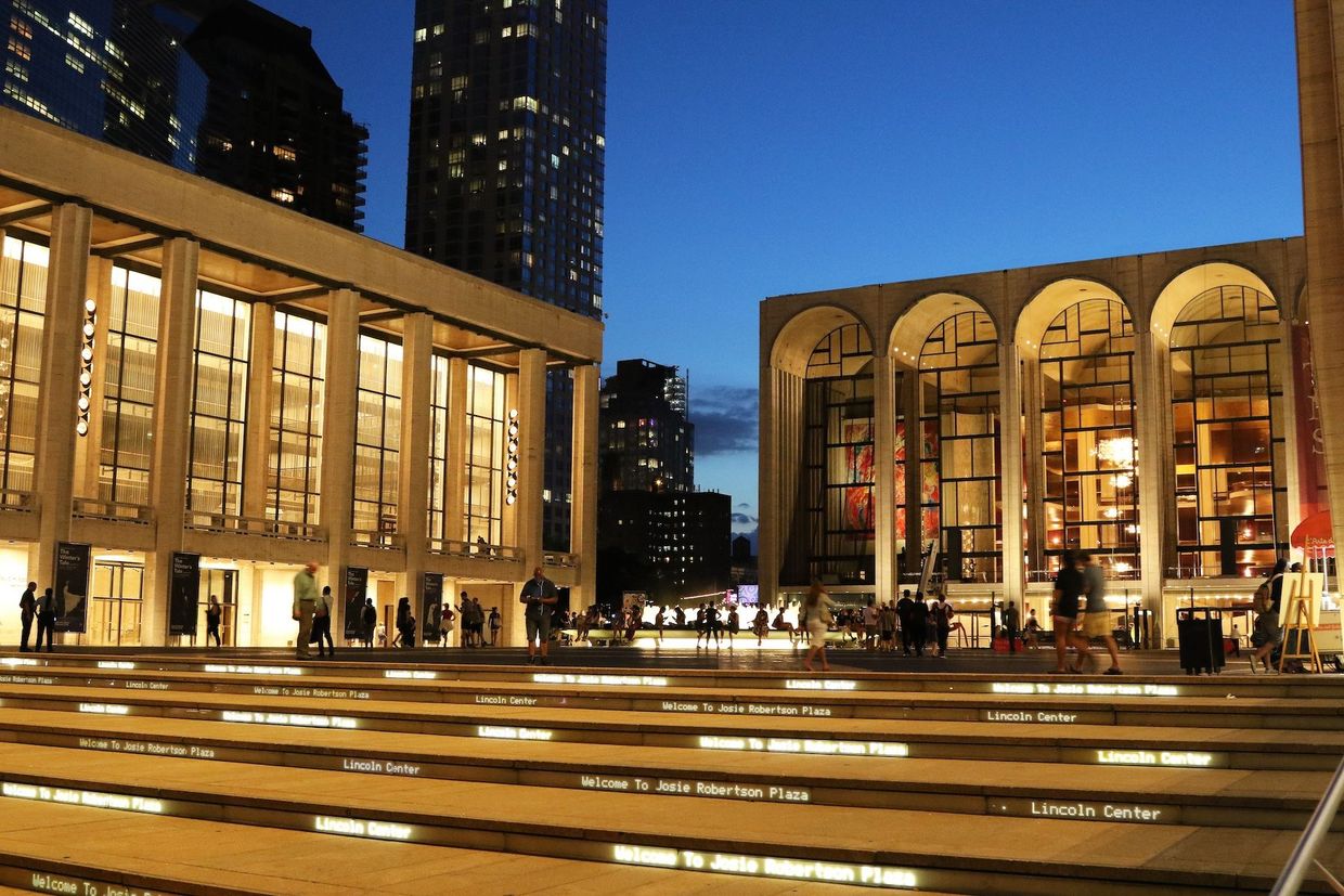 Lincoln Center at dusk. Large buildings with lights, people walking.