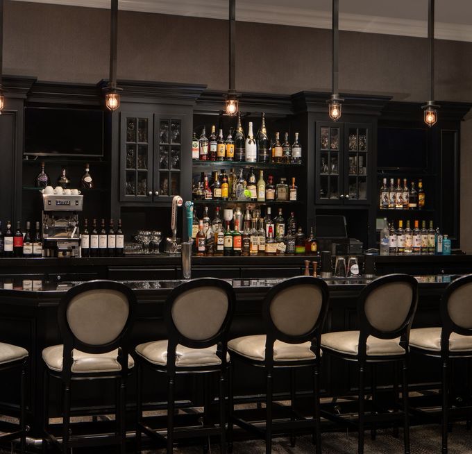 Bar with black cabinetry, liquor bottles, and high-backed chairs. Overhead lights illuminate the space.