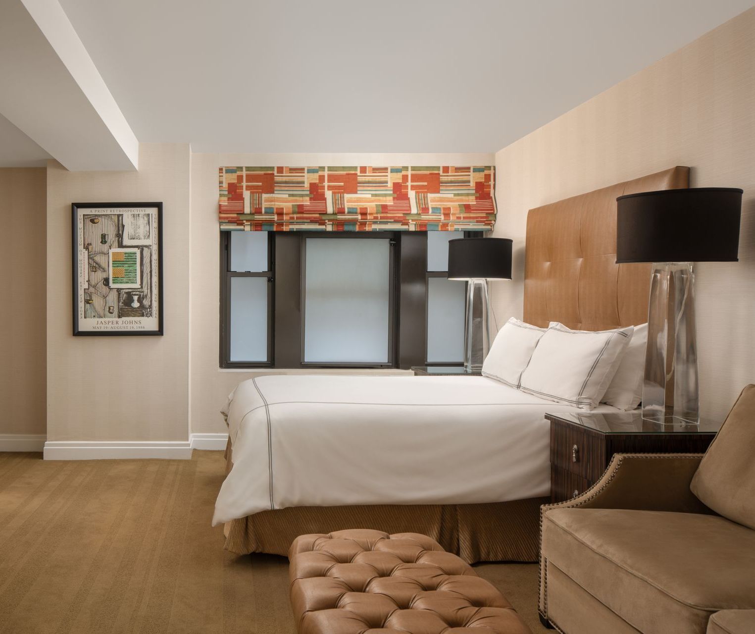 Modern hotel room with bed, tufted bench, sofa, and large windows with gray blinds and patterned valance