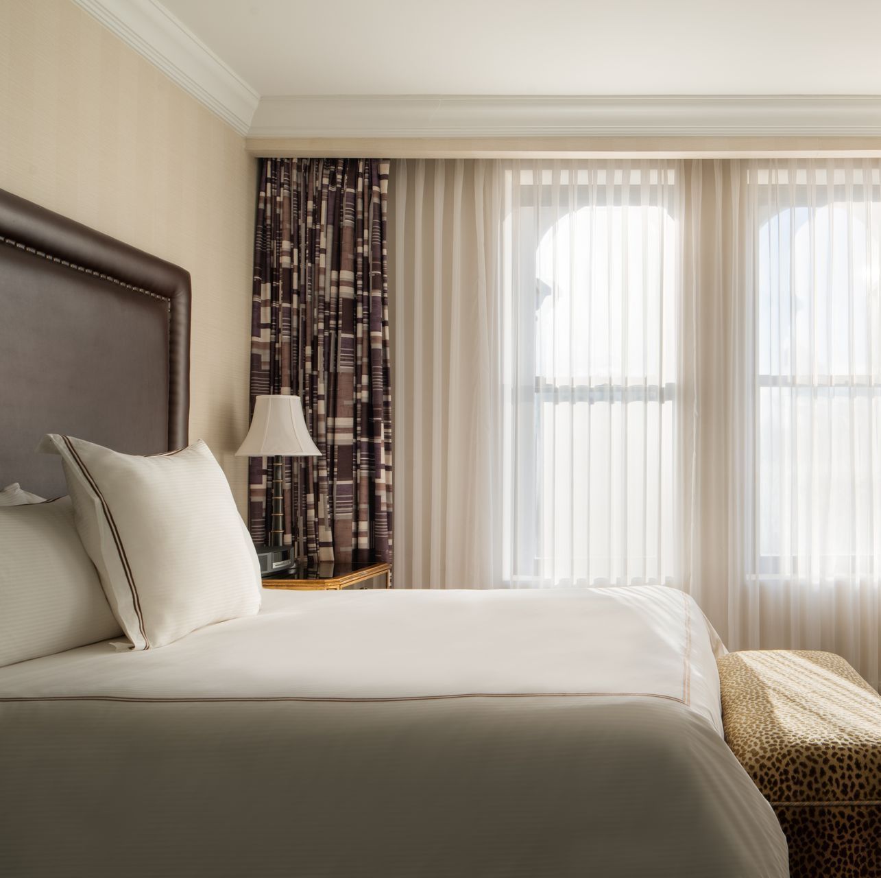 Bright hotel bedroom with a neatly made bed, lamp, and sheer curtains over tall windows.