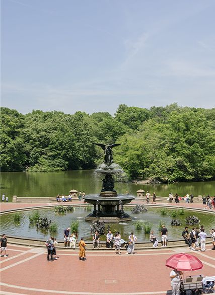 People strolling around a fountain in a park with trees and a pond beyond.