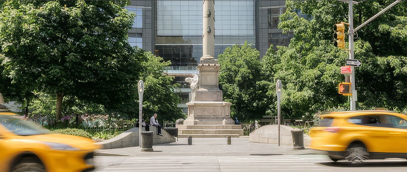 Yellow taxis pass a stone monument in front of a glass building and trees
